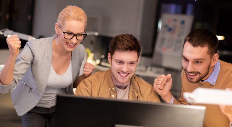 business, success and teamwork concept - happy coworkers with computer celebrating victory at night office