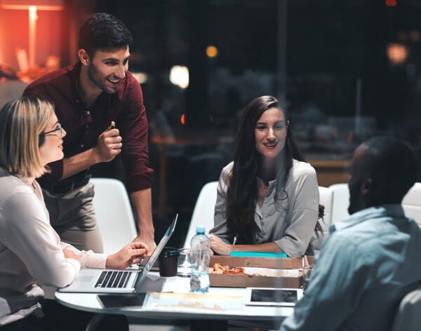 Business people, laptop and discussion for project at night in office, creative agency and overtime food for design. Teamwork, support and problem solving in pizza meeting for deadline and planning.